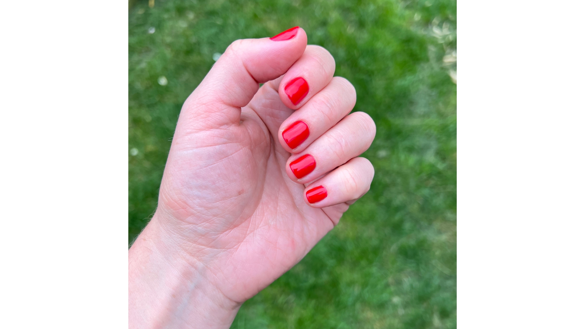 A close-up of Digital Beauty Editor, Fiona McKim's hand, featuring a bright red manicure, pictured above a grassy background