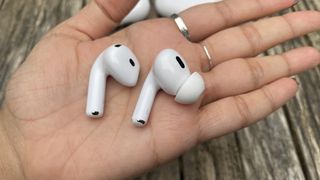 One earbud each from the Apple AirPods Pro 2 and AirPods 4 in the palm of someone's hand in front of a wooden surface.