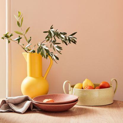 A vibrant yellow vase against a blush pink backdrop next to a fruit bowl