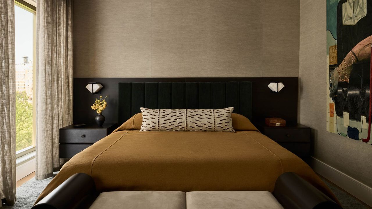 A bedroom with an extended headboard with matching dark wood side tables, white wall sconces, a mustard duvet with a patterned white bolster pillow with a white bench featuring a wood frame
