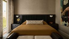 A bedroom with an extended headboard with matching dark wood side tables, white wall sconces, a mustard duvet with a patterned white bolster pillow with a white bench featuring a wood frame