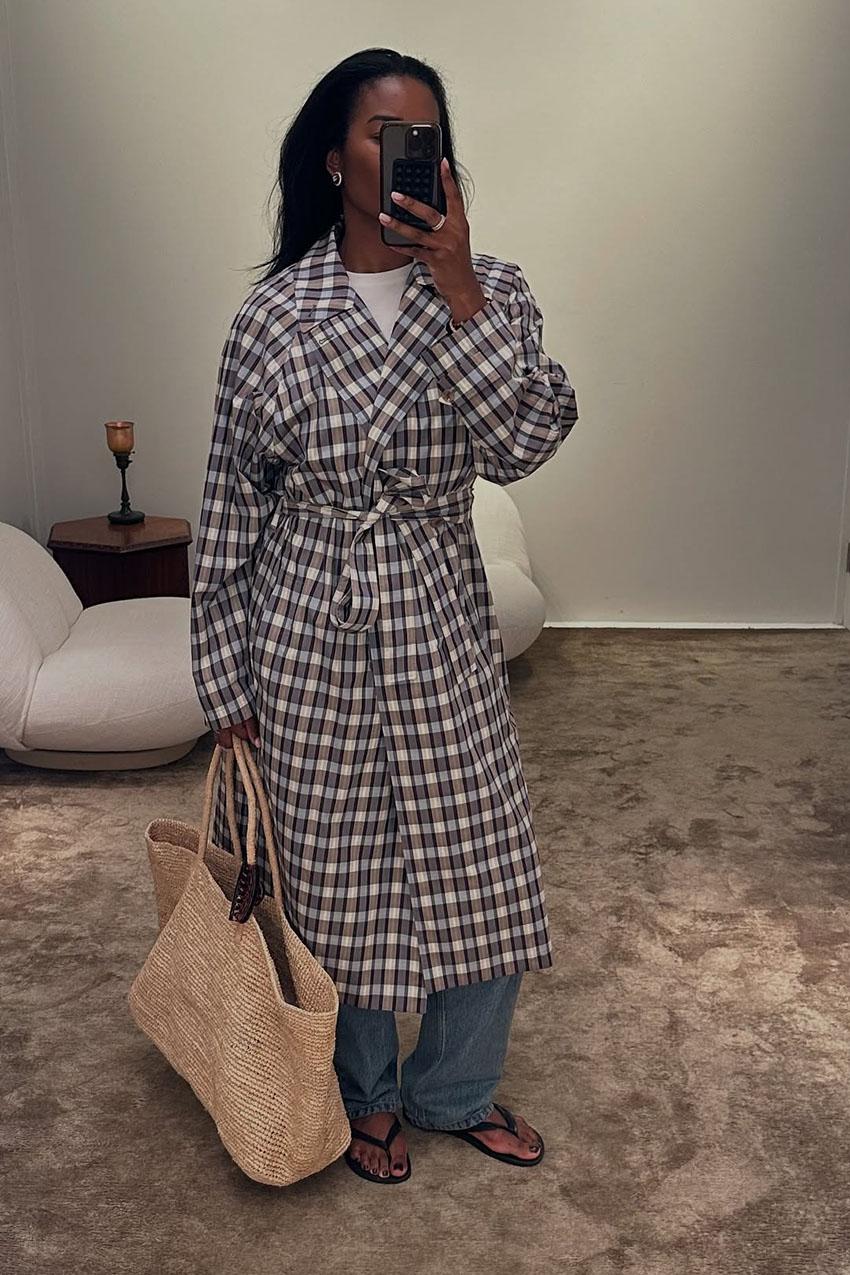 A woman wearing a plaid trench coat, blue jeans, and thong sandals