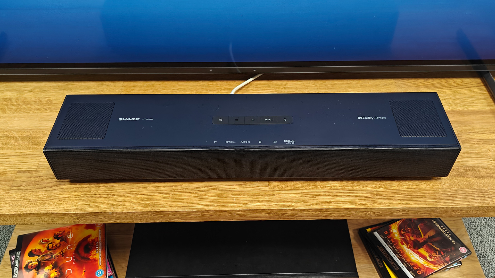 Sharp HT-SB700 Dolby Atmos soundbar on wooden AV rack in front of TV with Blu-ray discs seen on lower shefl