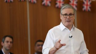 UK Prime Minister Keir Starmer delivers a speech in front of brown wall draped in Union Flags.