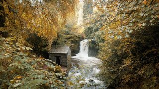 A small brick building next to a river, with a waterfall in the background and golden leaves in the foreground.