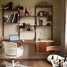 A beige-painted home office with a wall-mounted desk and storage unit in dark wood paired with a white rattan desk chair