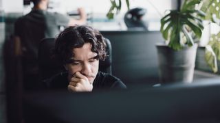 Male IT worker sitting at a desk in an open plan office space looking stressed and worried.