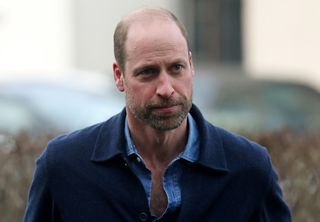 Prince William wearing an unbuttoned denim shirt and a blue jacket standing outside with a serious expression