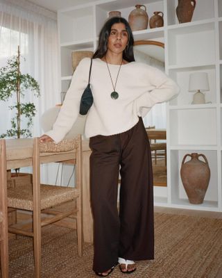 British woman wears flowy brown The Row pants and an oversized sweater.