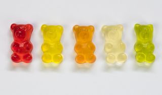 A close up showing a line of gummy bear sweets on a white background. (L-R) red, yellow, orange, clear and green gummy bears.