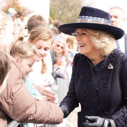 Queen Camilla wearing a blue coat and hat shaking hands with people