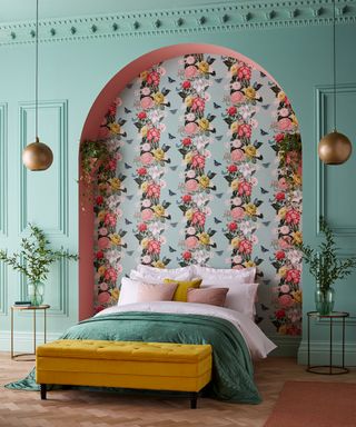 Colorful floral wallpaper in a pale mint green bedroom with wall panelling, a teal-dressed bed and mustard yellow stool.