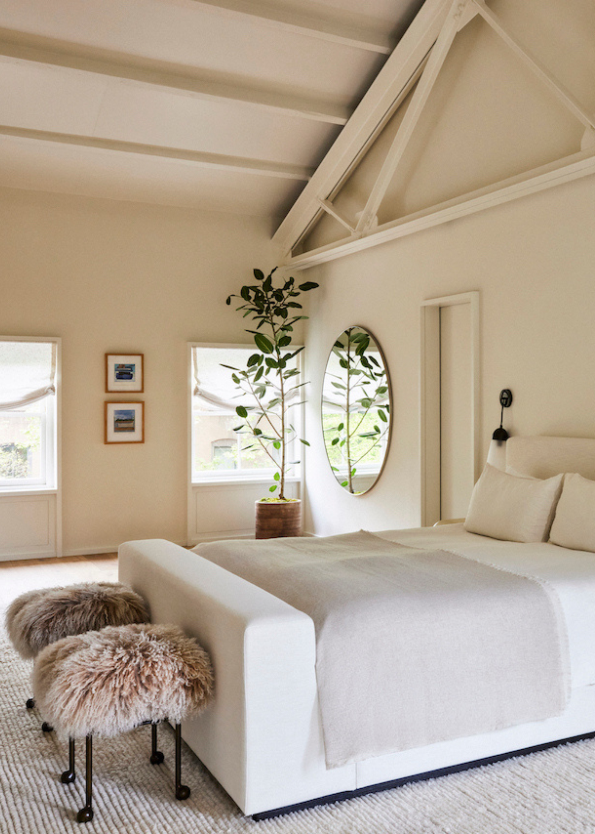 A cream bedroom with a vaulted ceiling, a large white bed with fur stools at the foot of the bed and a potted plant near a round mirror