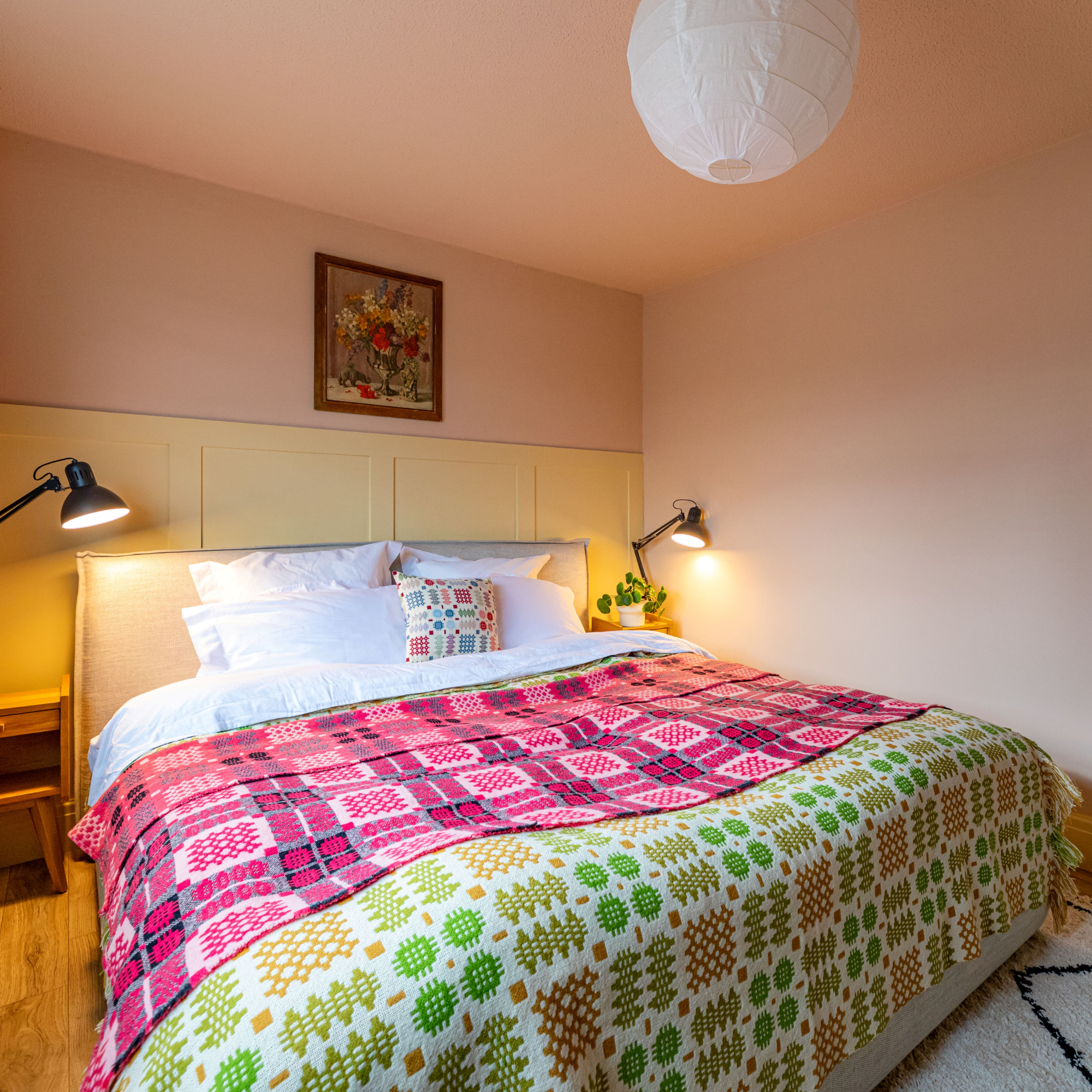 Bedroom with double bed and colourful blankets, bedside tables and lamps, green curtains