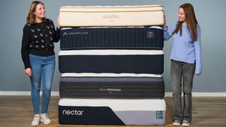 Two members of the Tom's Guide sleep team smile at each other while standing either side of a stack of the best mattresses we've tested in 2026, including Saatva, DreamCloud, Helix and Nectar