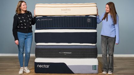 Two members of the Tom's Guide sleep team smile at each other while standing either side of a stack of the best mattresses we've tested in 2026, including Saatva, DreamCloud, Helix and Nectar