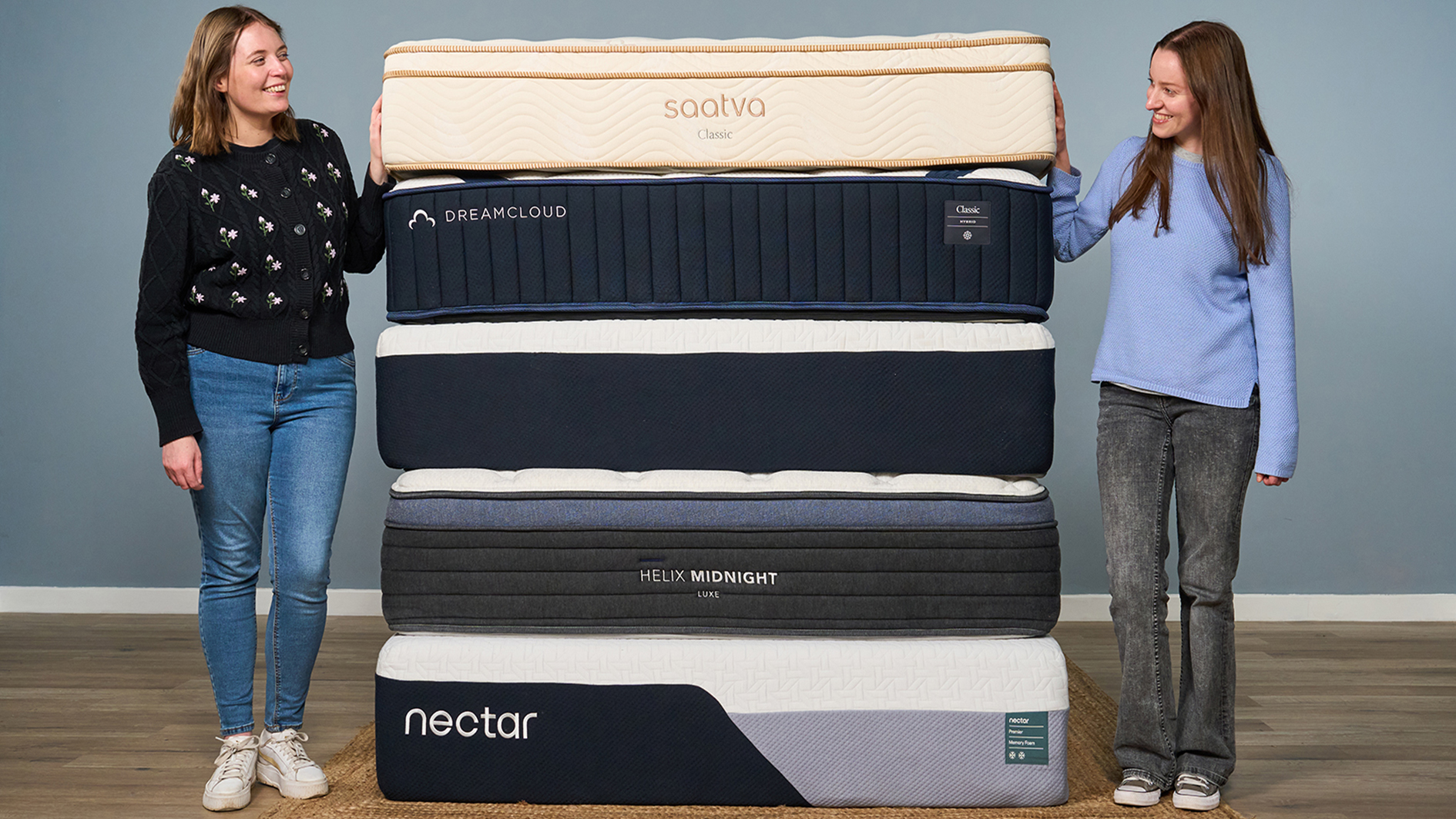 Two members of the Tom's Guide sleep team smile at each other while standing either side of a stack of the best mattresses we've tested in 2026, including Saatva, DreamCloud, Helix and Nectar