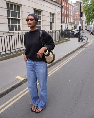 Marilyn wears blue straight leg jeans, black sandals, a black sweater, black sunglasses, a black headband and a straw shopping bag.