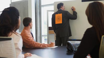 A man in a suit writes on a white board in a work meeting. There is a piece of paper with a target drawn on it taped to his back.