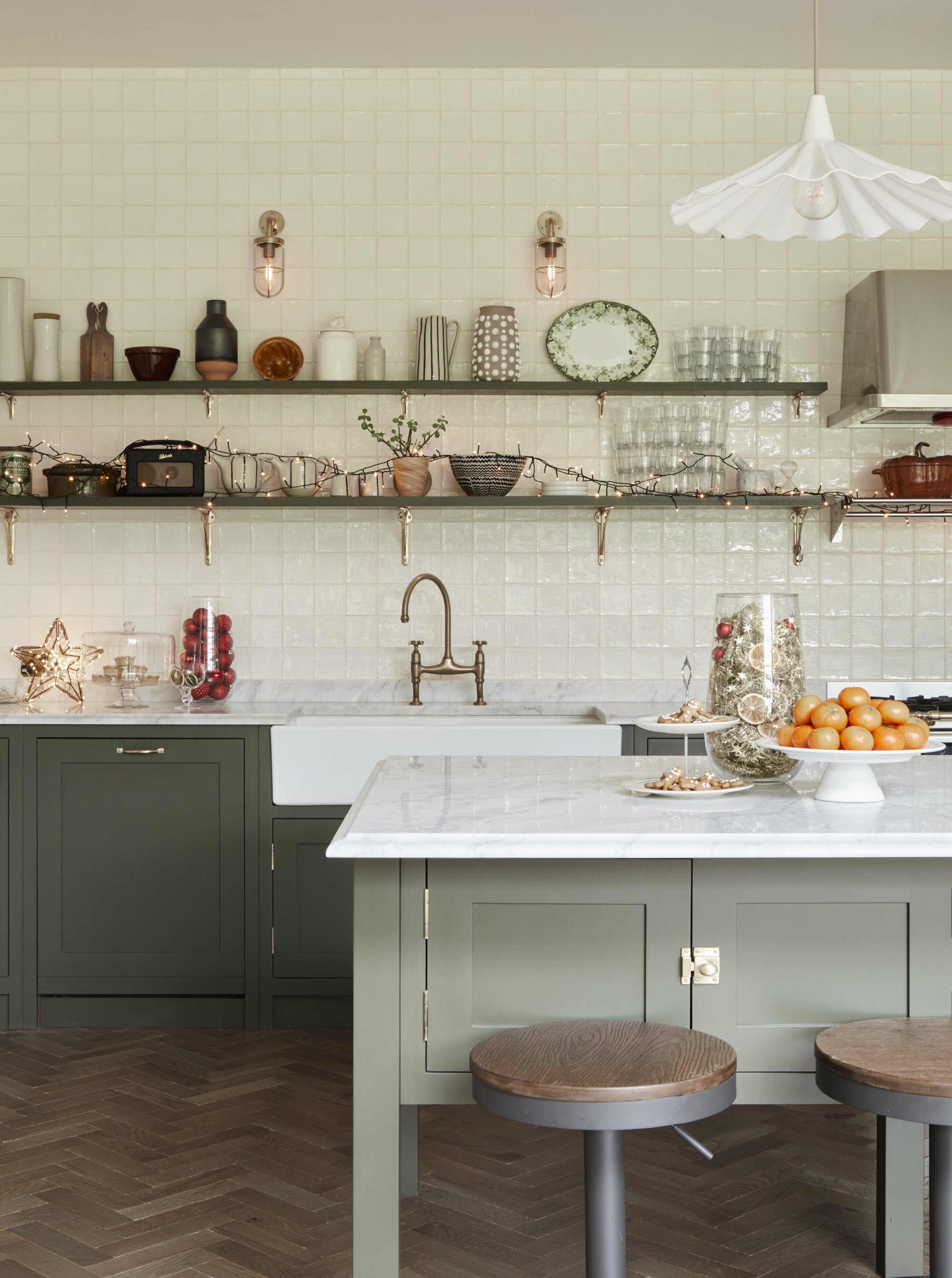 A kitchen with sage green inset cabinetry, two bar stools at a kitchen island, and Christmas decorations on the open shelving