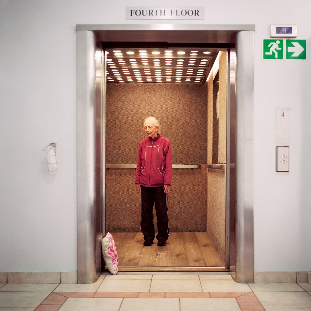 An elderly person stands in an open lift wearing a pink fleece