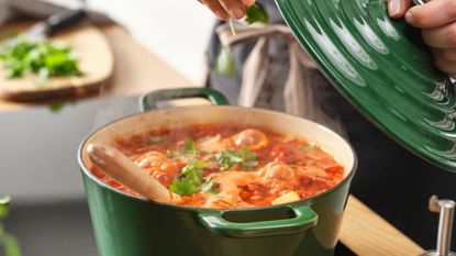 A dark green Dutch oven on a stove with someone lifting the lid off. A bubbling tomato pasta inside. 