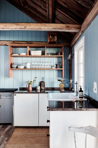 A kitchen with pale blue shiplap walls, wooden beams and vaulted wooden ceiling with white cabinetry and dark grey granite worktop