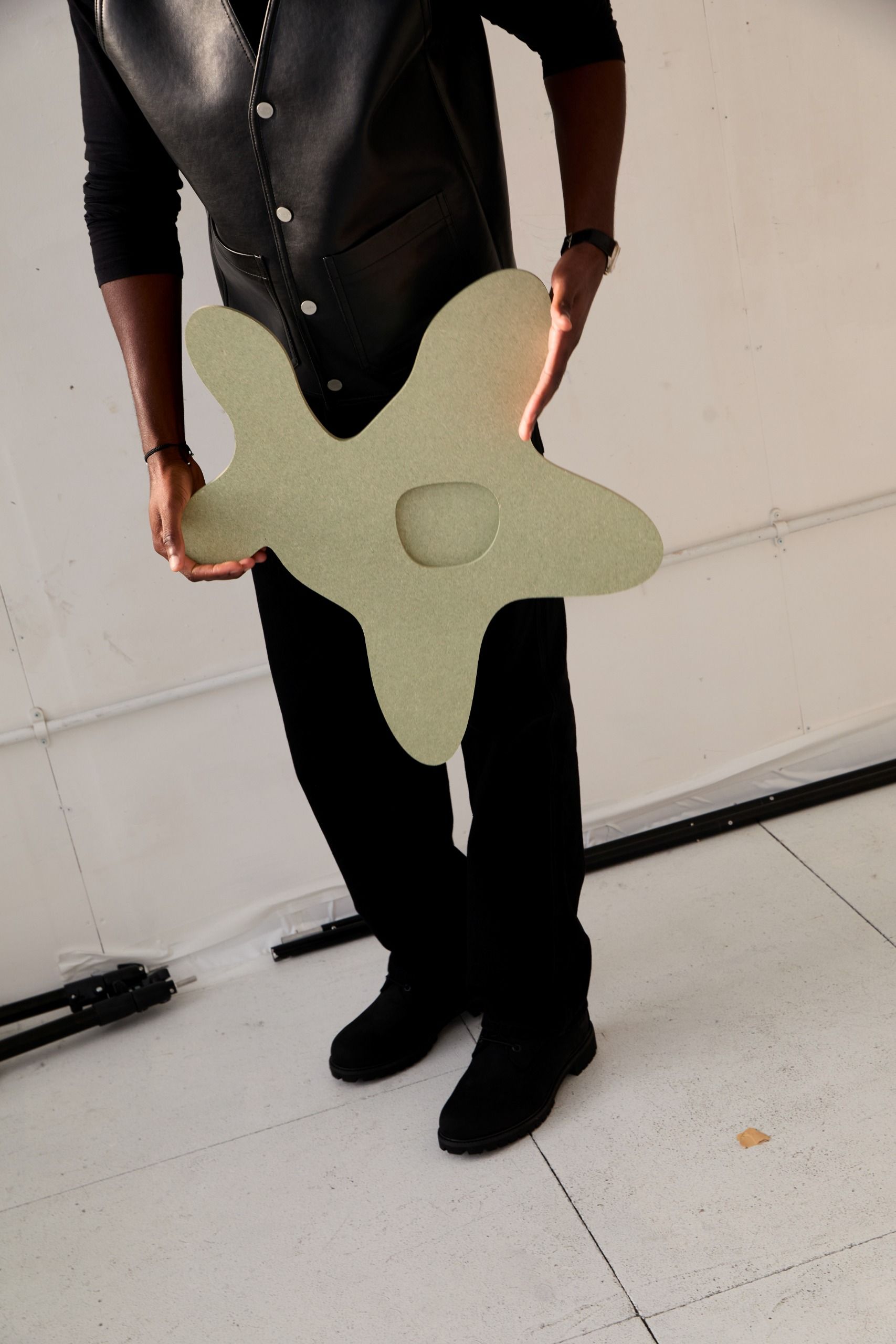 A young man poses in his industrial studio dressed in casual smartwear, sitting or standing in front of a gray wall with his sculptural creations.