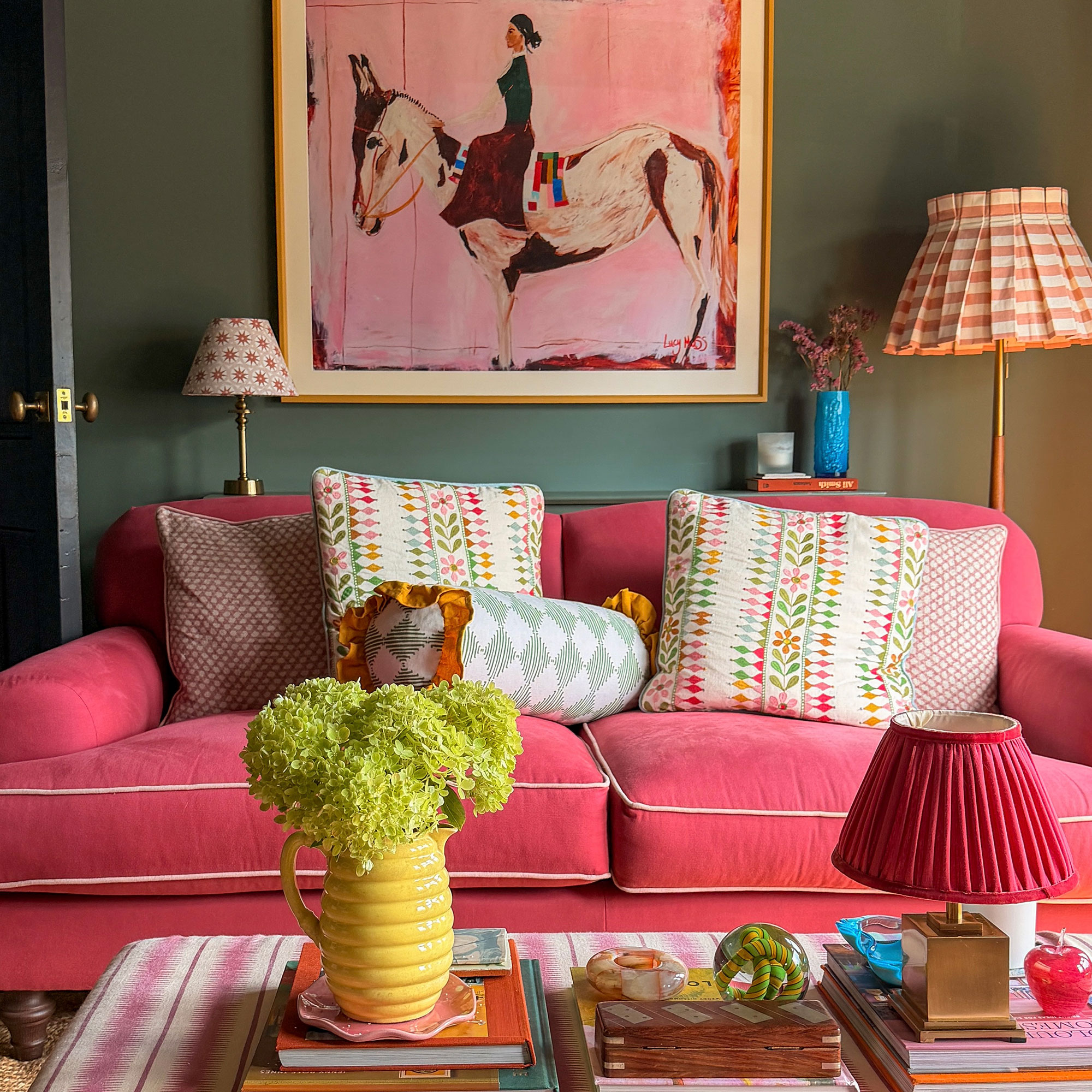 Green living room with pink sofa, colourful patterned cushions, horse artwork on wall and patterned ottoman coffee table with coffee table books, jug of flowers and wireless table lamp
