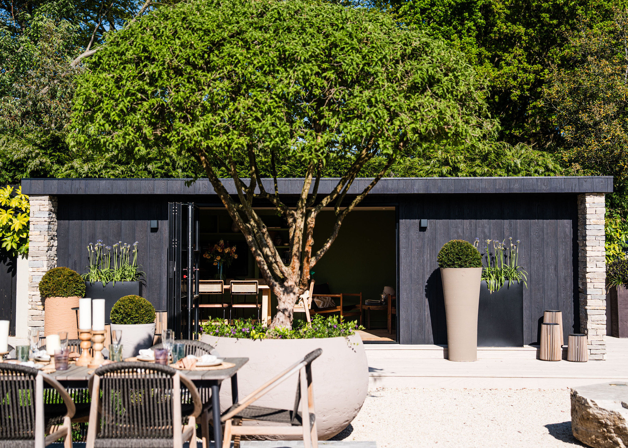 a charred timber clad garden room with open folding glass doors. There is also a large dining table in the garden with candles on top and tableware.