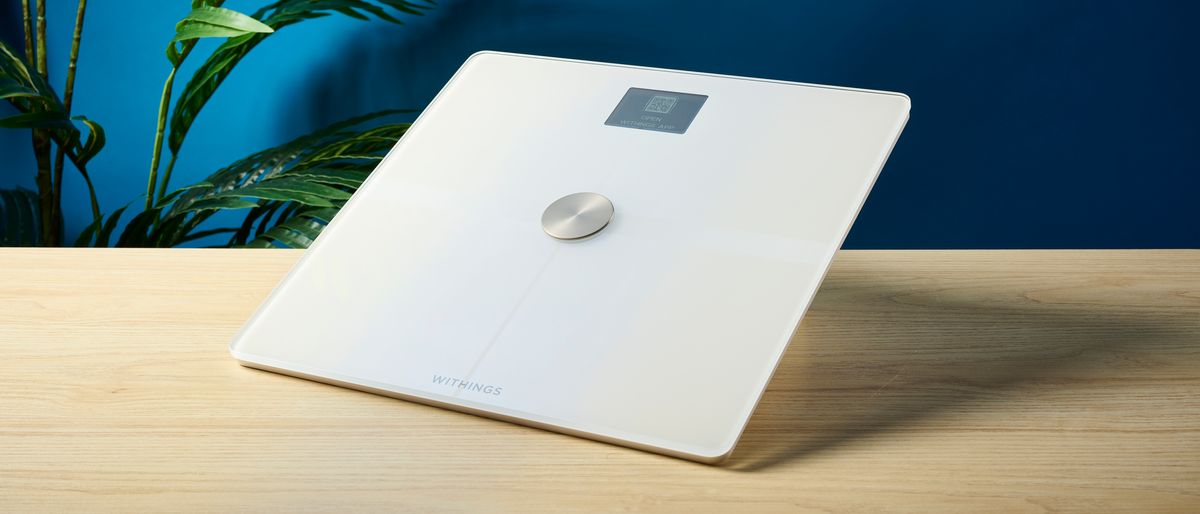 a white smart scale by withings rests on a wooden table, showing its LED screen and weighing scales