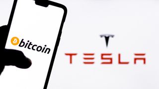 Somebody using a smartphone with Bitcoin on the face in front of a massive Tesla logo