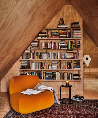 A triangle-shaped wooden room with open book storage and a orange lounge chair