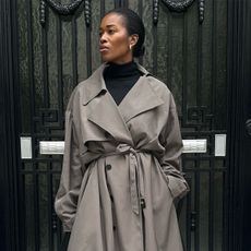 Woman standing in front of black wall, wearing black turtleneck and grey trench coat.