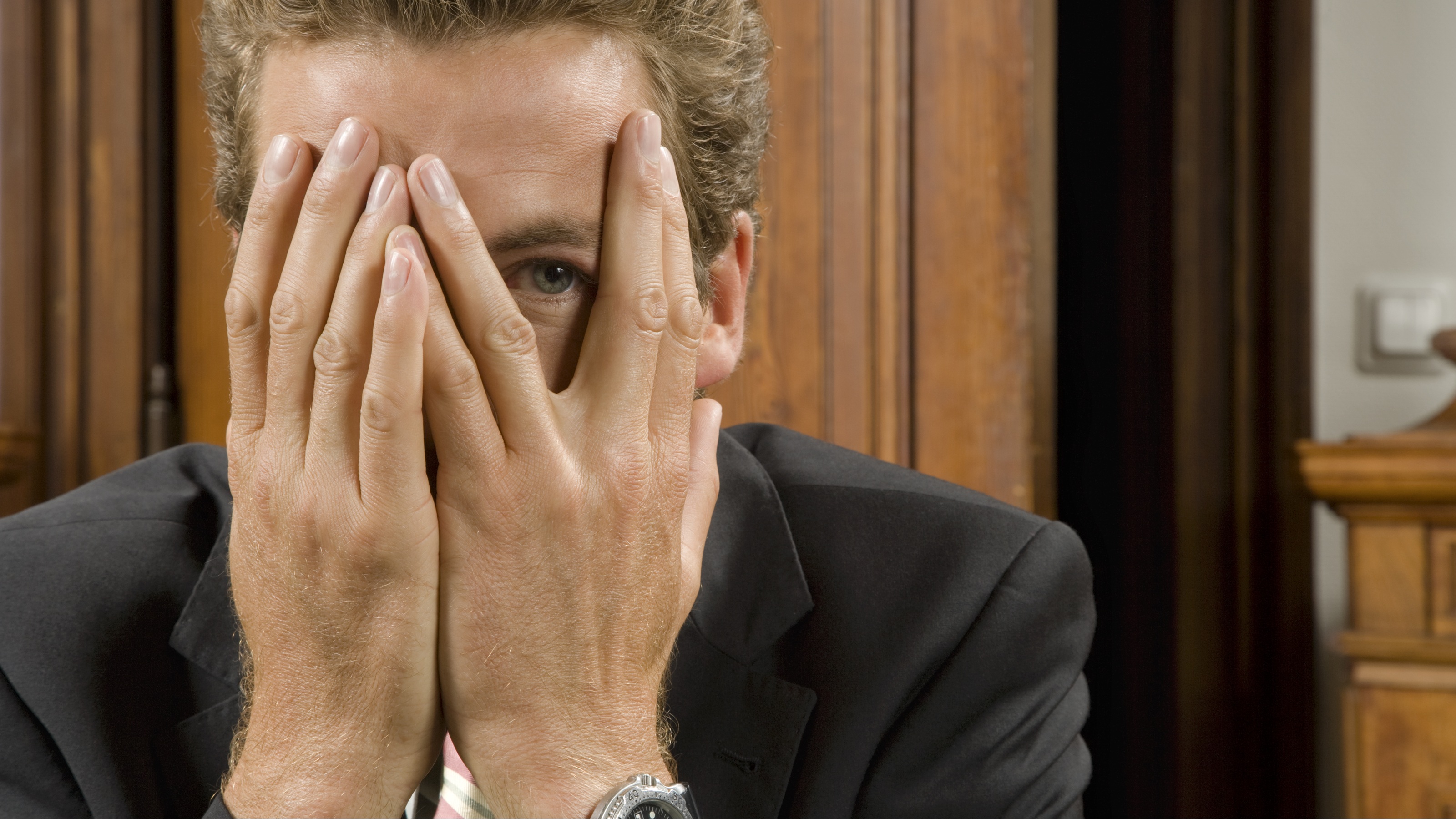 A businessman looks out with one eye from behind his hands.