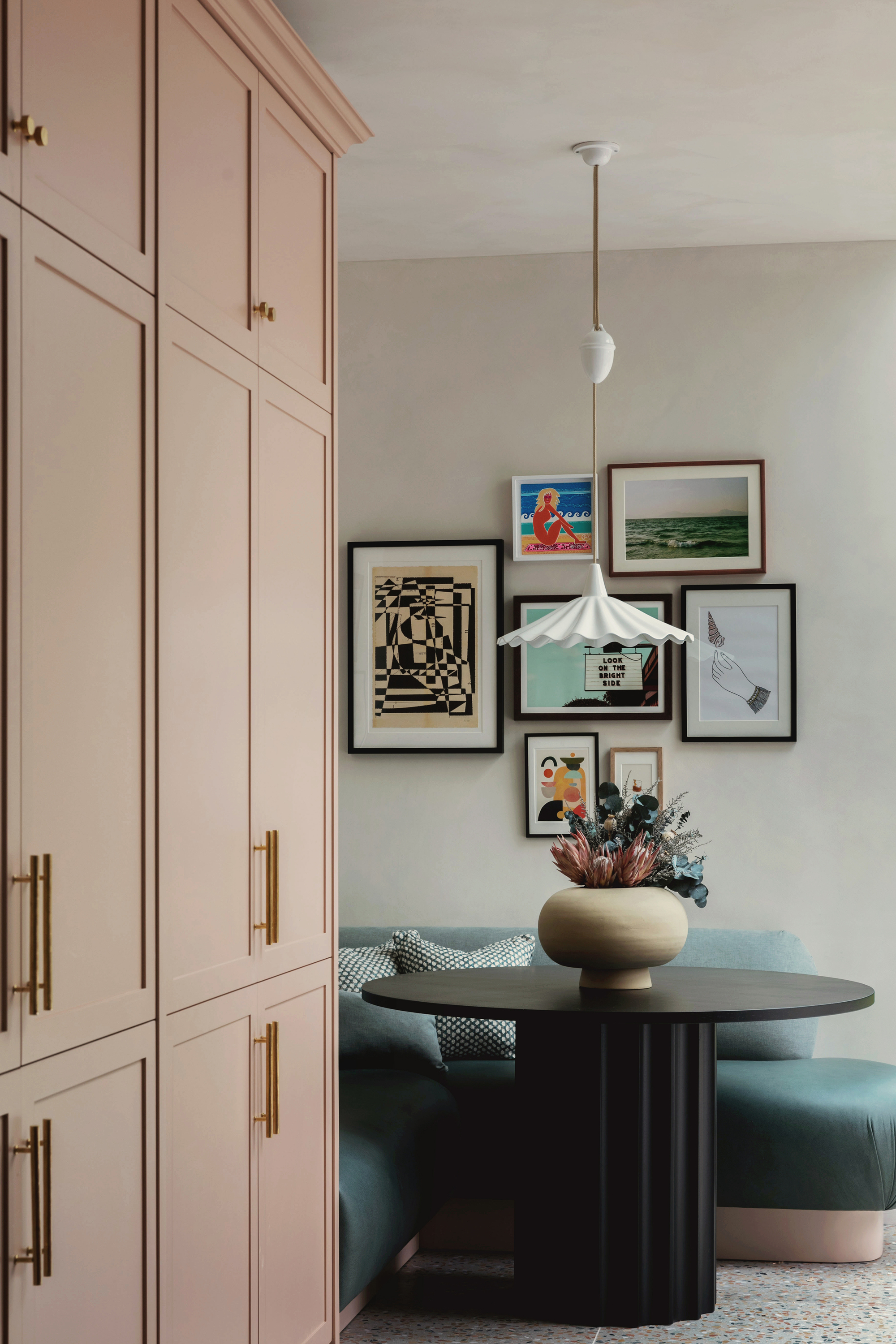banquette seat in corner of kitchen with small black round dining table, pendant light hanging above, gallery wall behind, vase of flowers on the table, and pink cabinets in the foreground