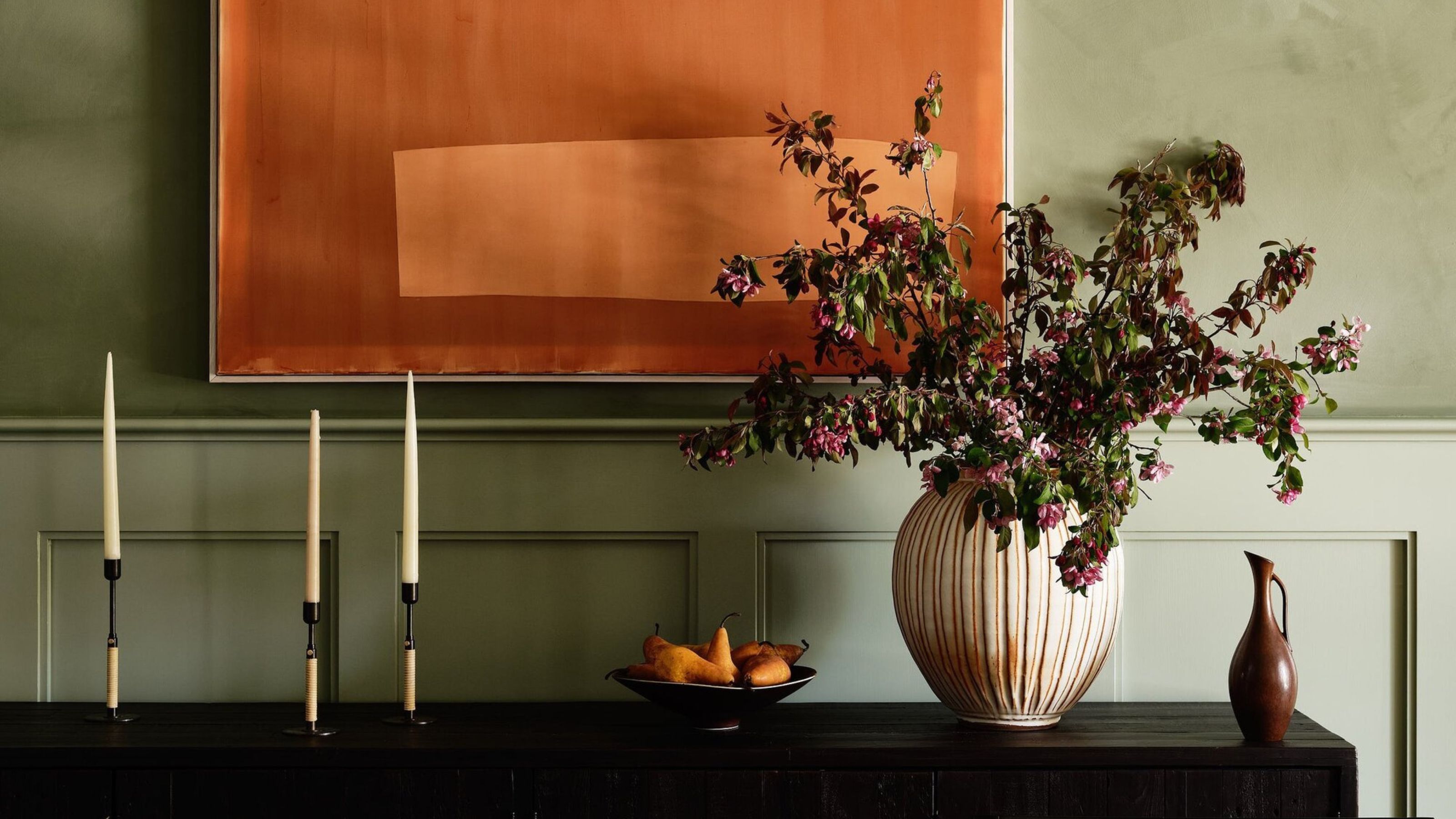 close up of table console with vase of flowers, bowl of fruit, and three candlesticks. the wall behind is painted a sage green and there is an orange artwork hanging above