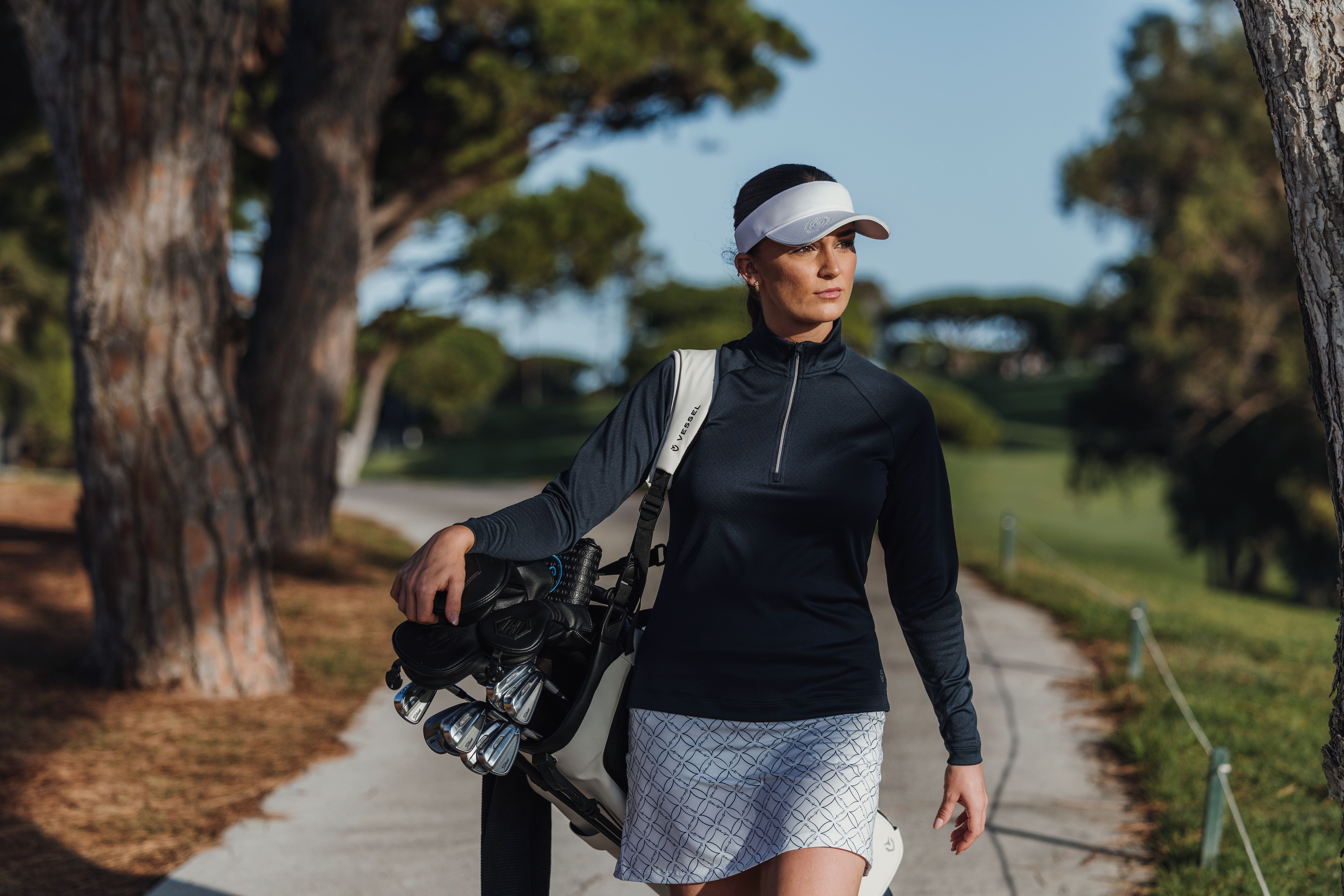 A female golfer carrying her bag wearing a visor, pullover and skirt