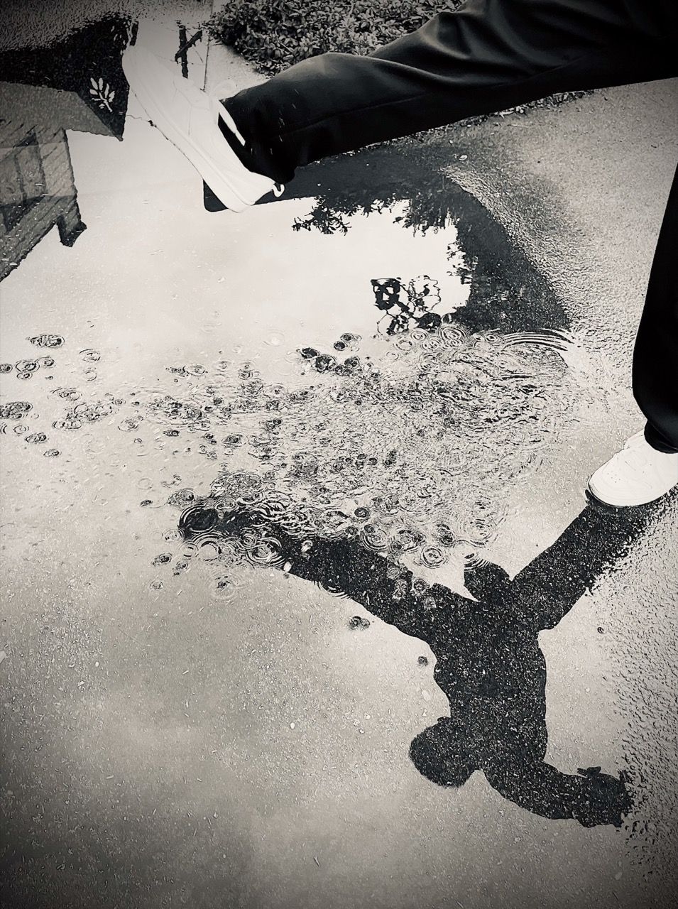 Photograph of a child's reflection in a puddle as they prepare to step over it, image is monochrome