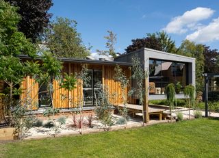 A tiny home with a large glazed window, timber cladding and shrubbery 