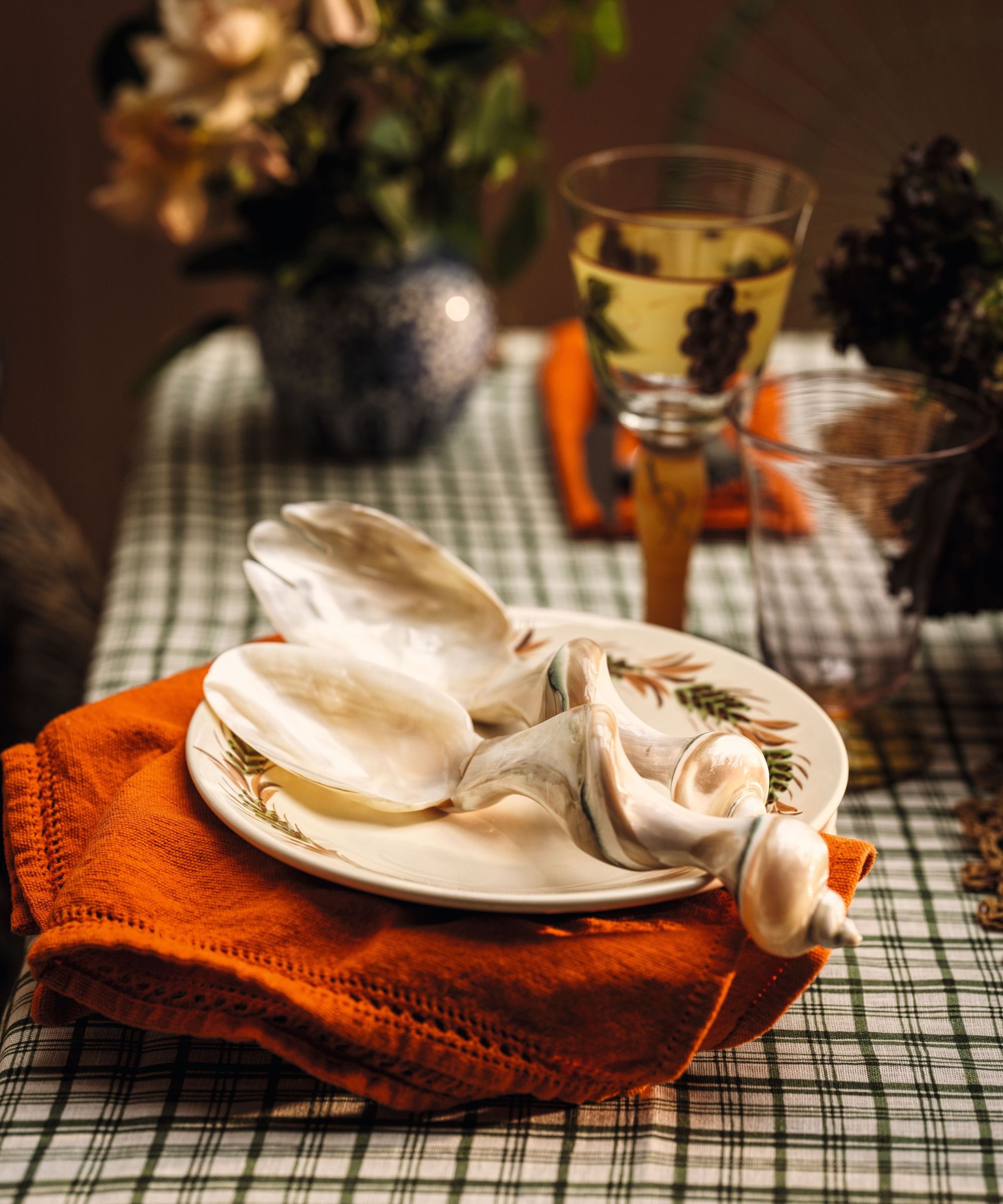 Mother-of-pearl salad servers rest on a patterned plate over a orange napkin and green-checked tablecloth with wine glasses in background.