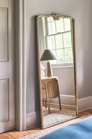 A large gold framed mirror with ornate details on the floor leaning against a wall