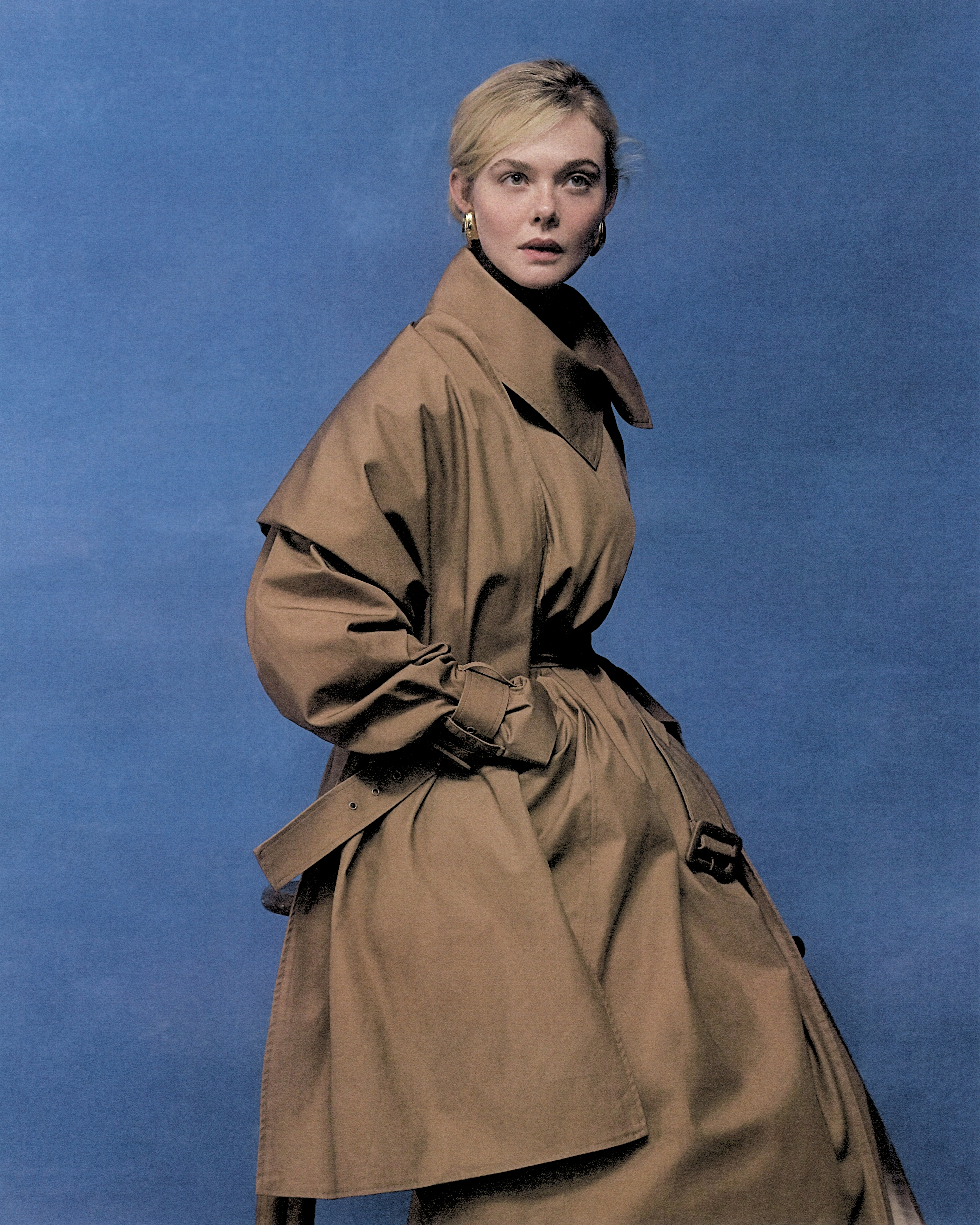 Elle Fanning shot by Szilveszter Mak&amp;oacute;. She is standing in front of a painted blue backdrop wearing a belted trench coat.