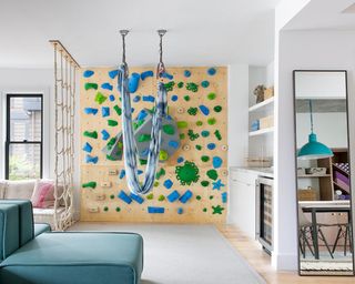 A living room with climbing wall made from wood and green/blue colored grips, hammock suspended from ceiling and rope climbing device