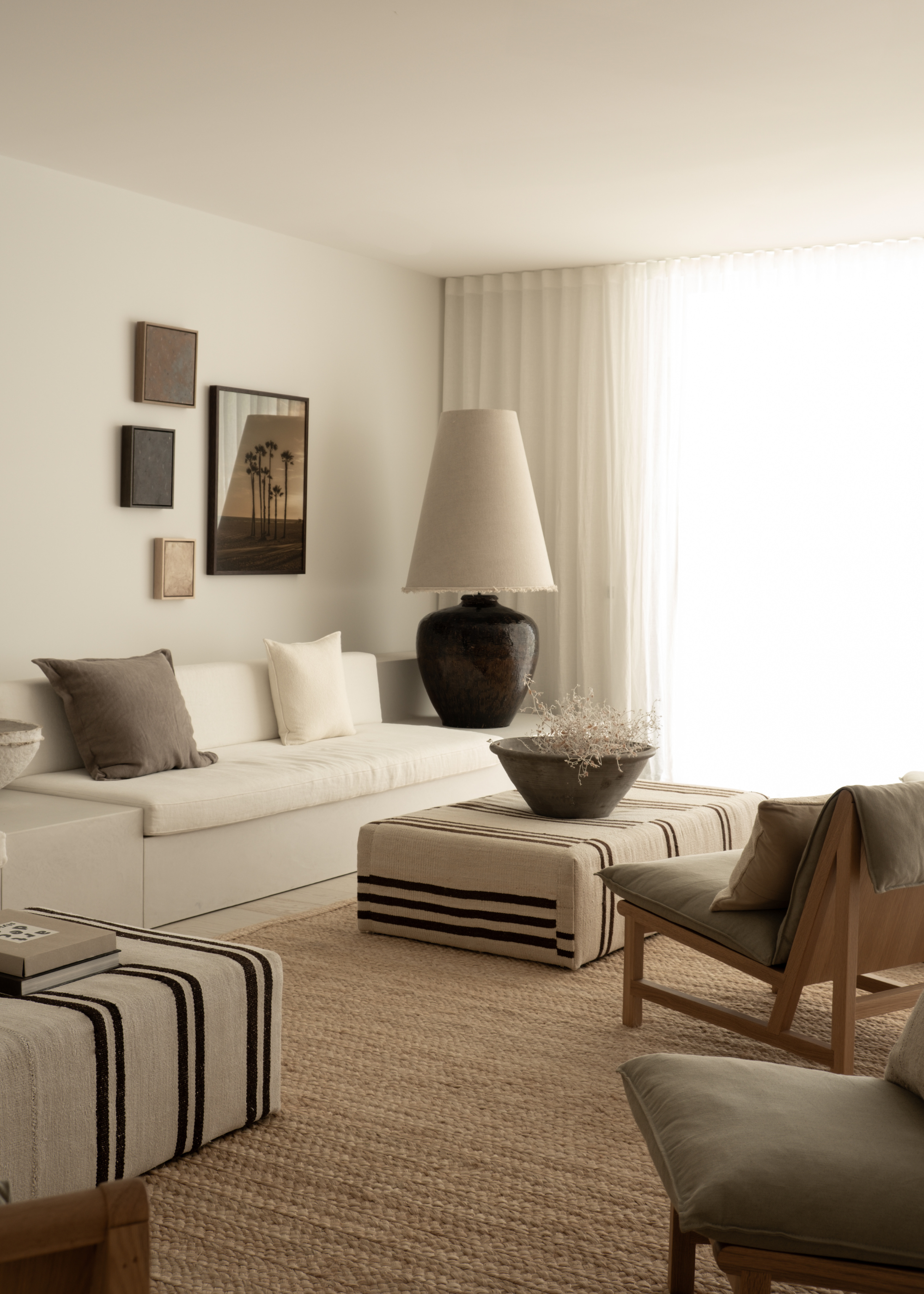 A minimalist living room with a white couch, a striped upholstered footstool, a gallery wall of prints and an oversized table lamp