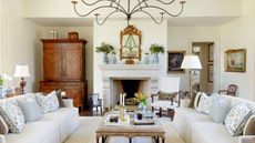Living room with fireplace, cream sofas, ottoman with antique decor, vintage paintings, mahogany dresser, and candelabra