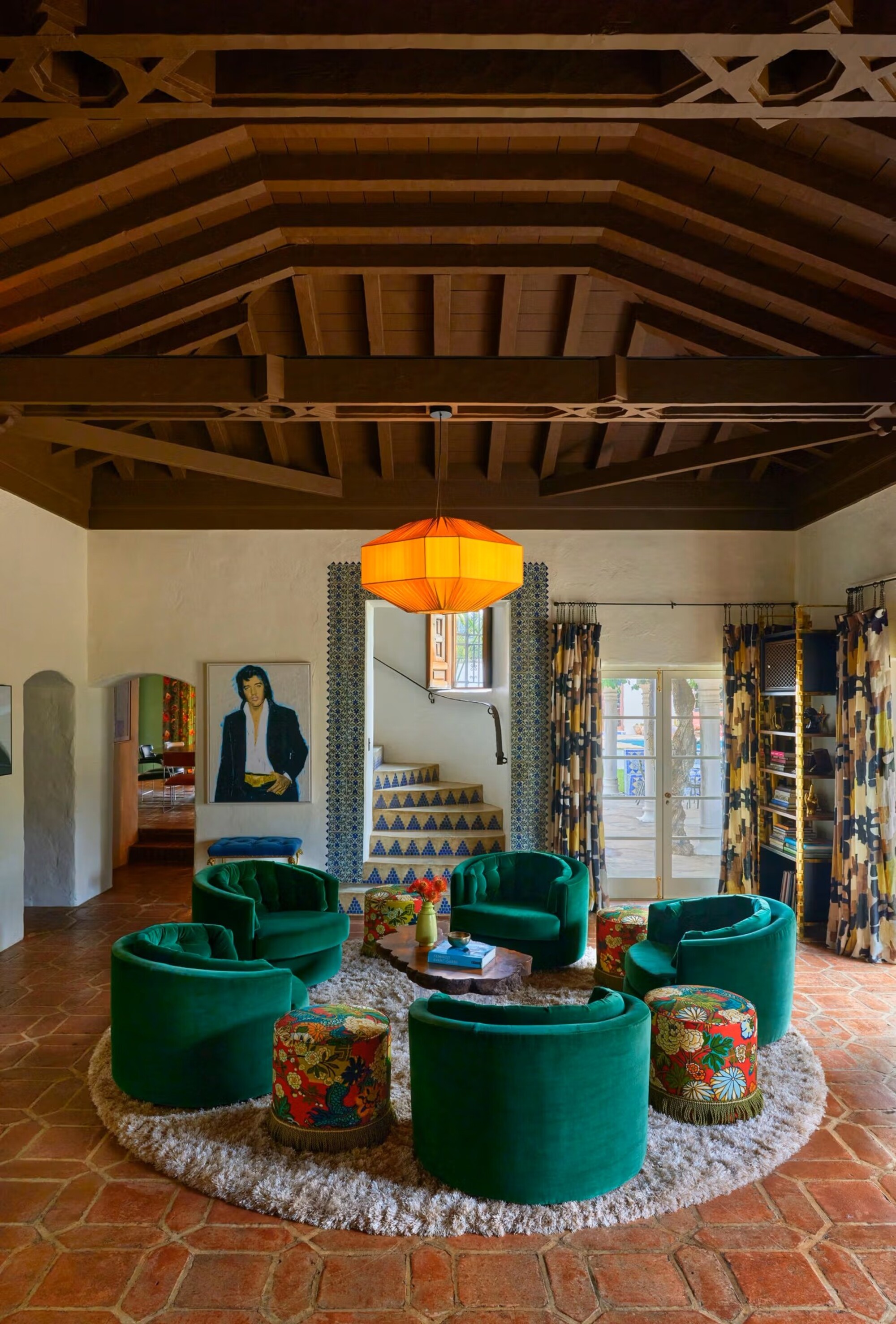 Image of a Spanish colonial living room with wooden ceilings and terracotta floors. There is a circle rug in the middle with an arrangement of teal accent chairs. A picture of Elvis is on the wall.
