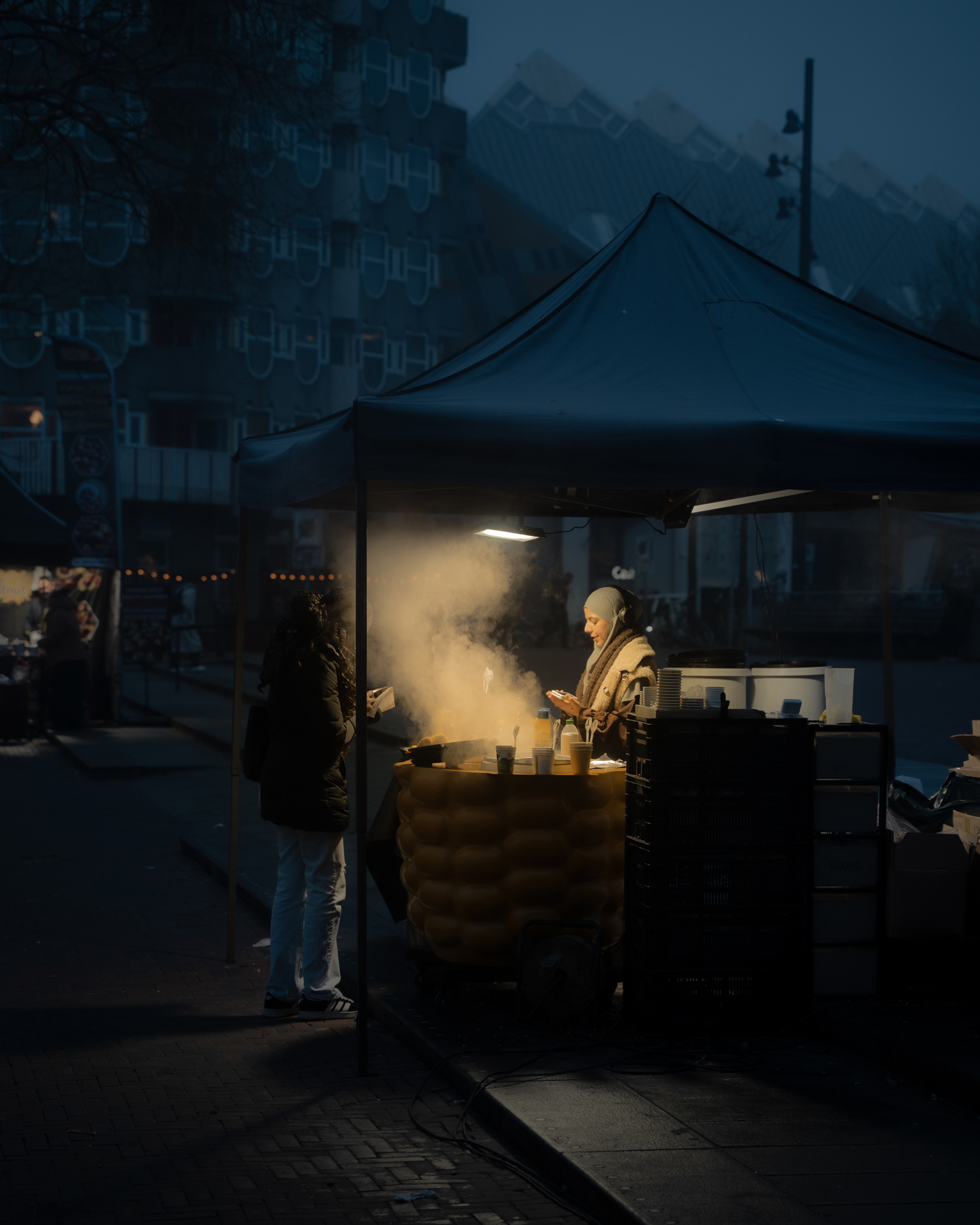 A night scene of a street food vendor under a tent, steaming food, and a customer standing nearby in a dimly lit urban area