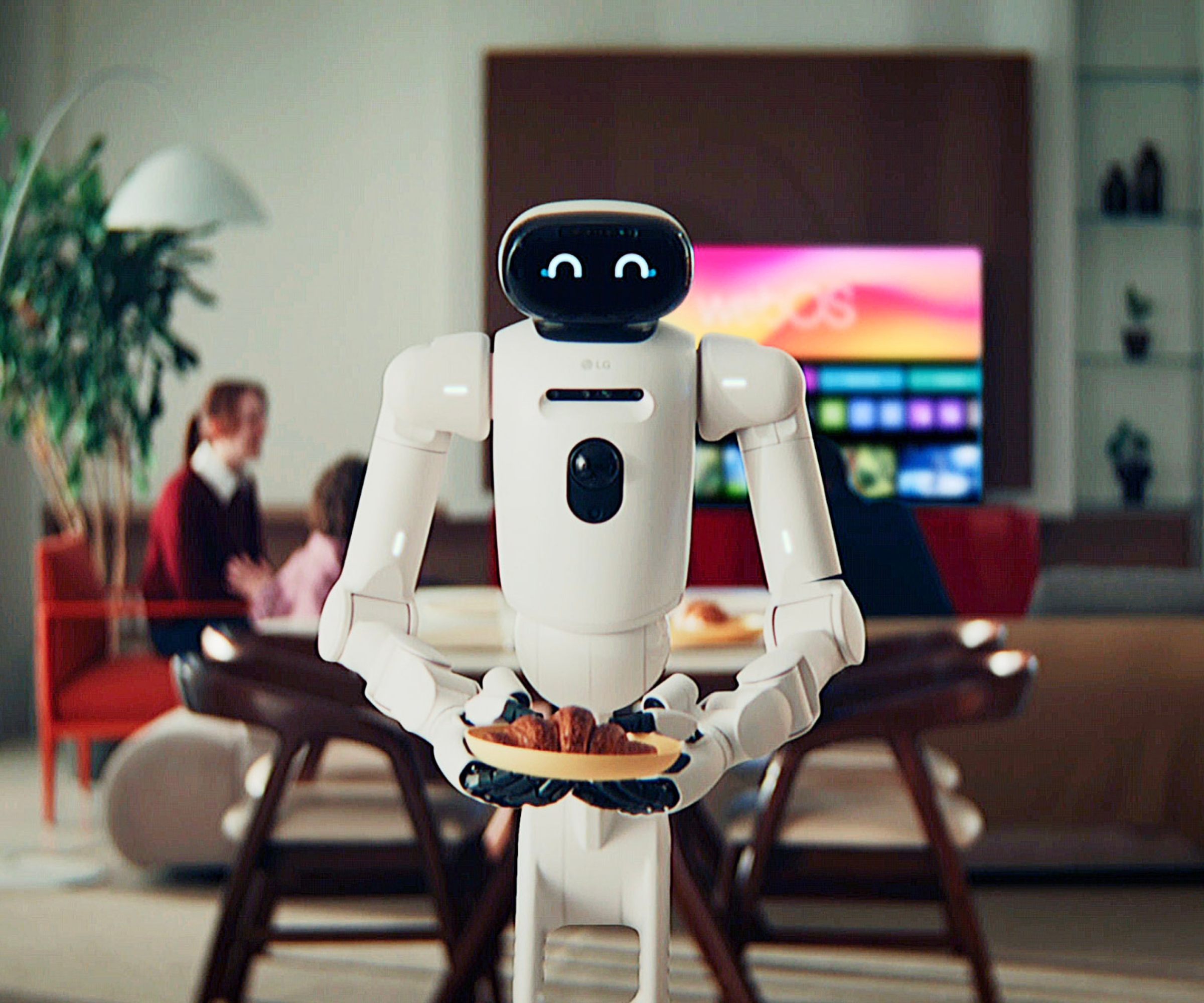 LG CLOiD holding croissant on small plate, pictured in front of family sat in living room around the TV.