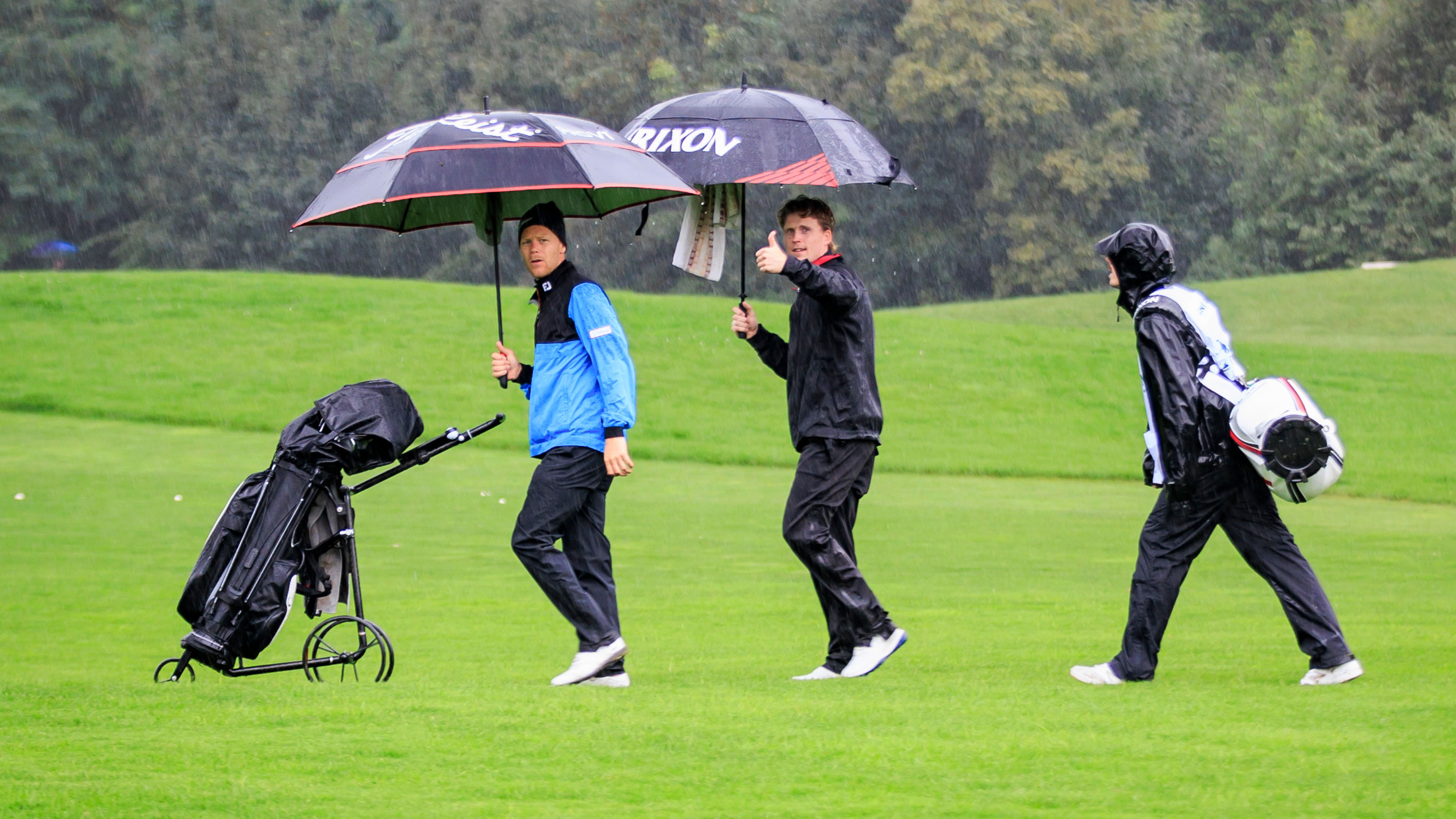 three golfers playing golf in the rain with their umbrellas up, with one of them giving a thumbs up to the camera